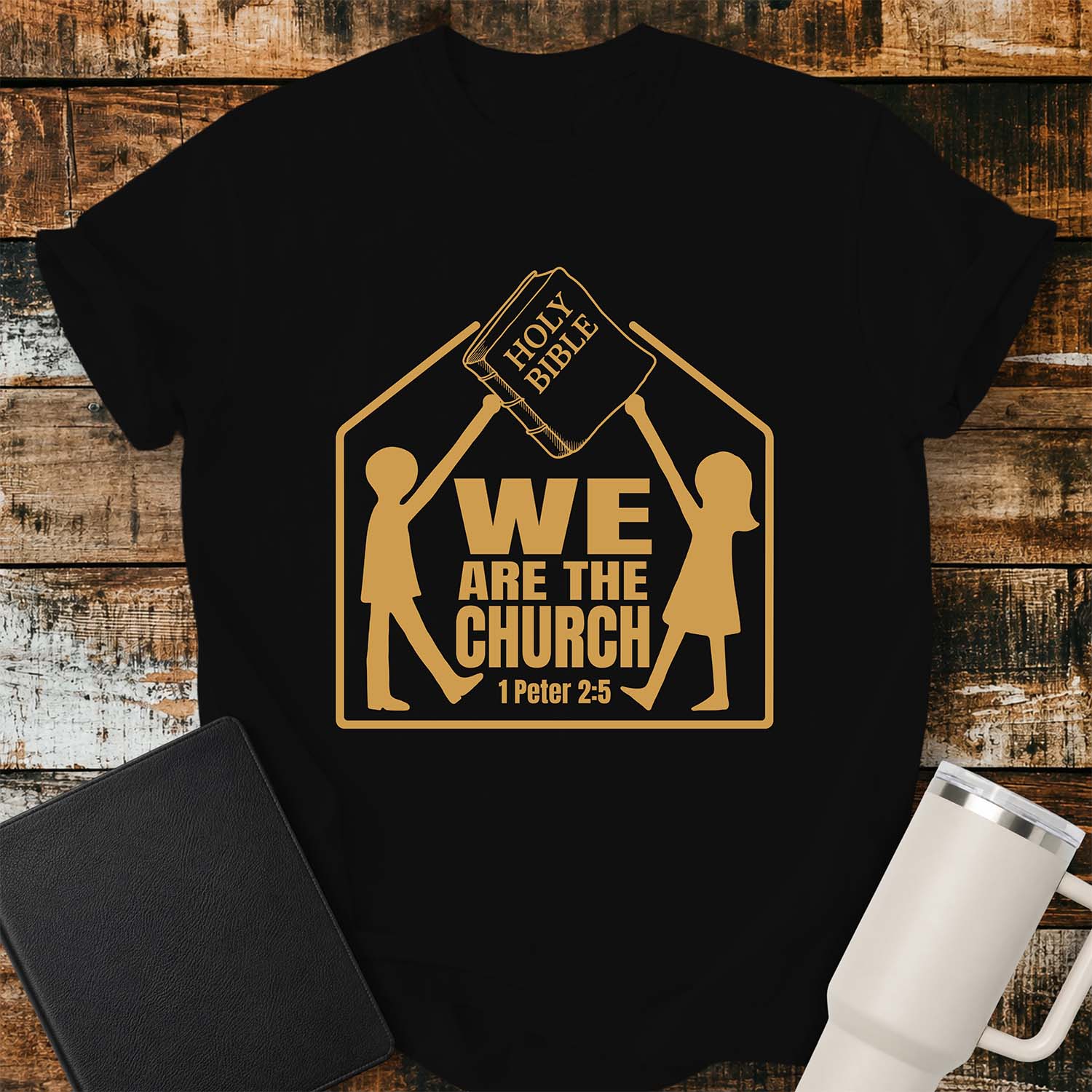Black T-shirt featuring a gold graphic of two children raising a Holy Bible above a house-shaped outline with the message “WE ARE THE CHURCH” and the Bible reference “1 Peter 2:5.” The shirt is displayed on a rustic wood plank background alongside a black Bible and a white insulated tumbler.