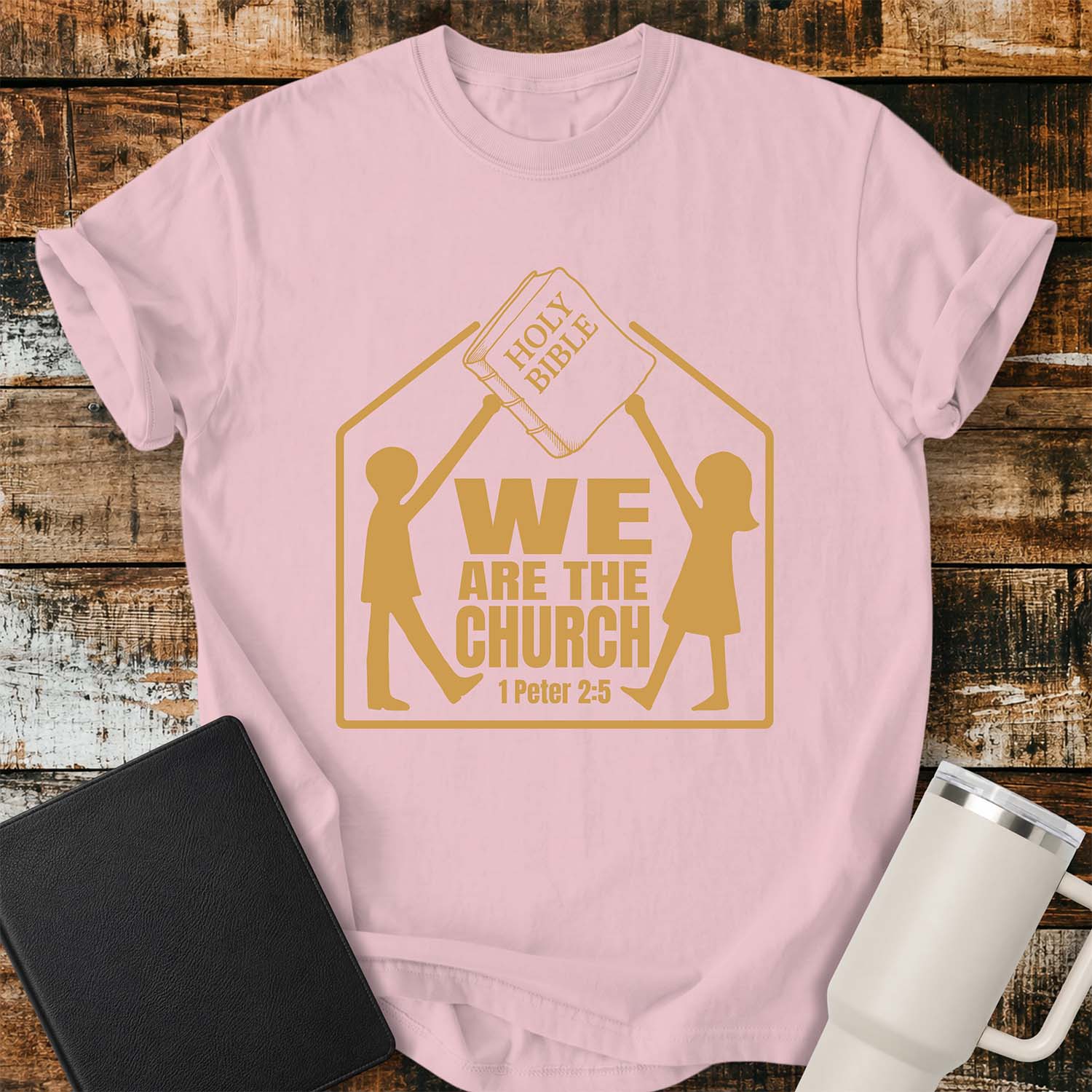 Blossom T-shirt featuring a gold graphic of two children raising a Holy Bible above a house-shaped outline with the message “WE ARE THE CHURCH” and the Bible reference “1 Peter 2:5.” The shirt is displayed on a rustic wood plank background alongside a black Bible and a white insulated tumbler.