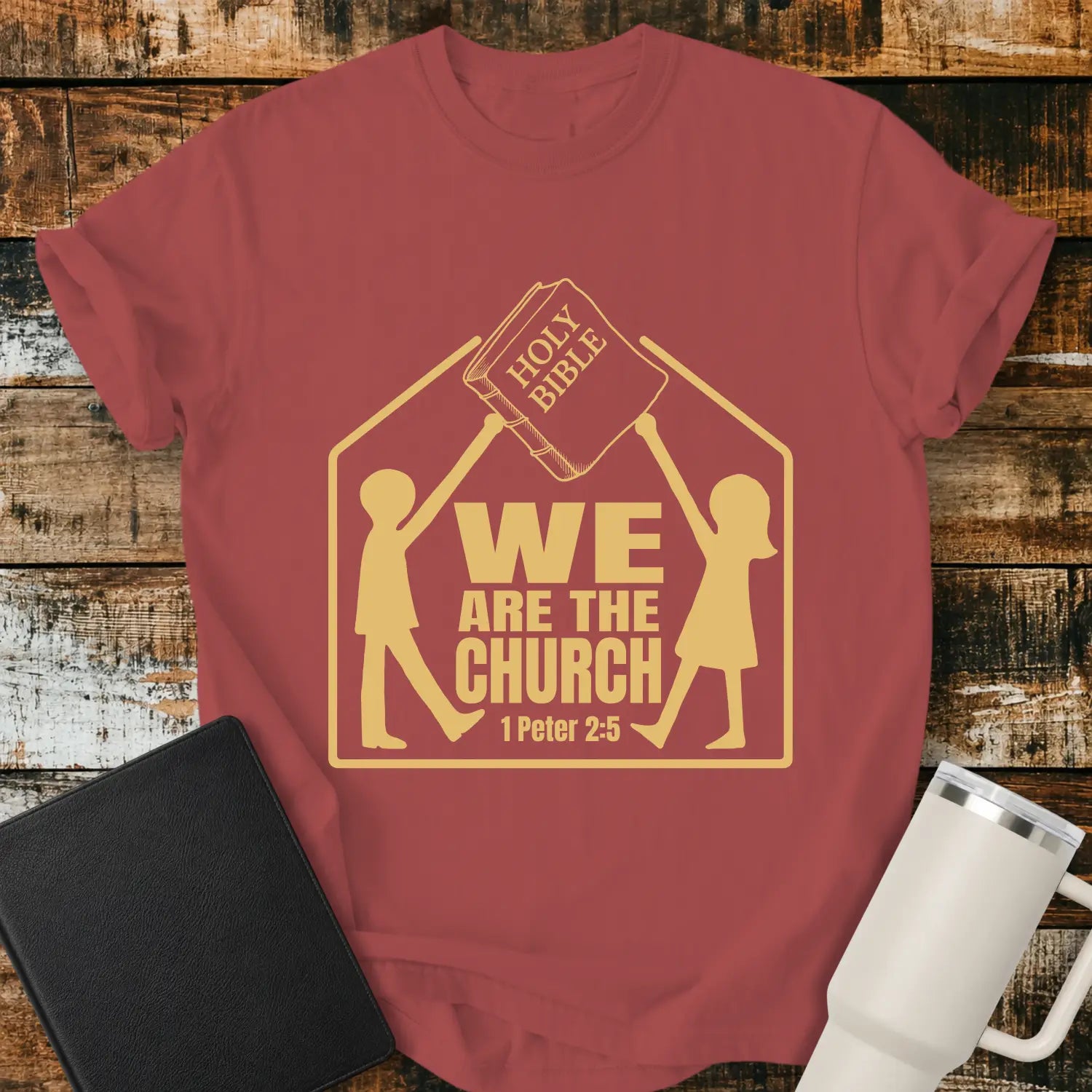 Crimson red graphic T-shirt mockup featuring the phrase “We Are the Church” with a 1 Peter 2:5 reference, showing two figures holding a Holy Bible above a church-shaped outline, styled with a black Bible and white tumbler on a rustic wood background.