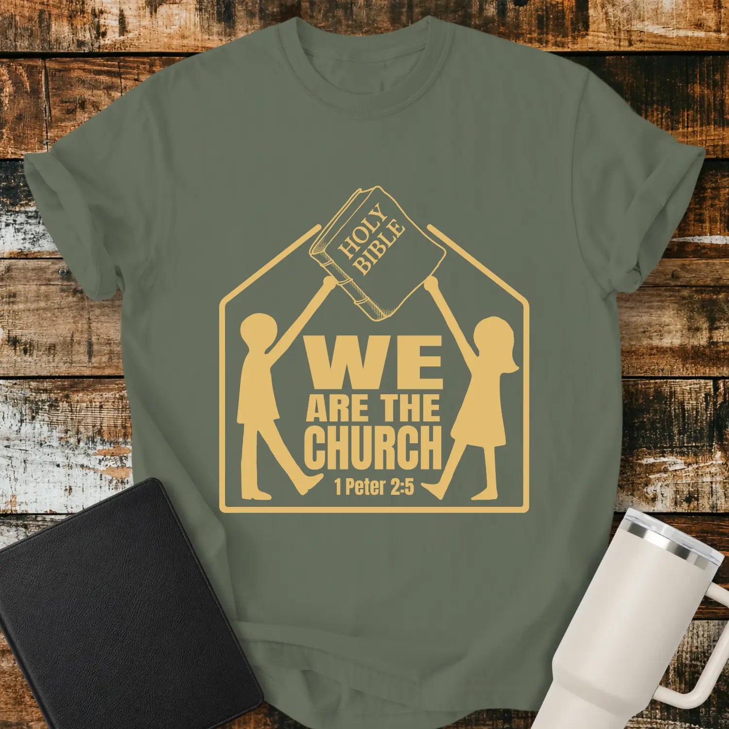moss green graphic T-shirt mockup featuring the phrase “We Are the Church” with a 1 Peter 2:5 reference, showing two figures holding a Holy Bible above a church-shaped outline, styled with a black Bible and white tumbler on a rustic wood background.