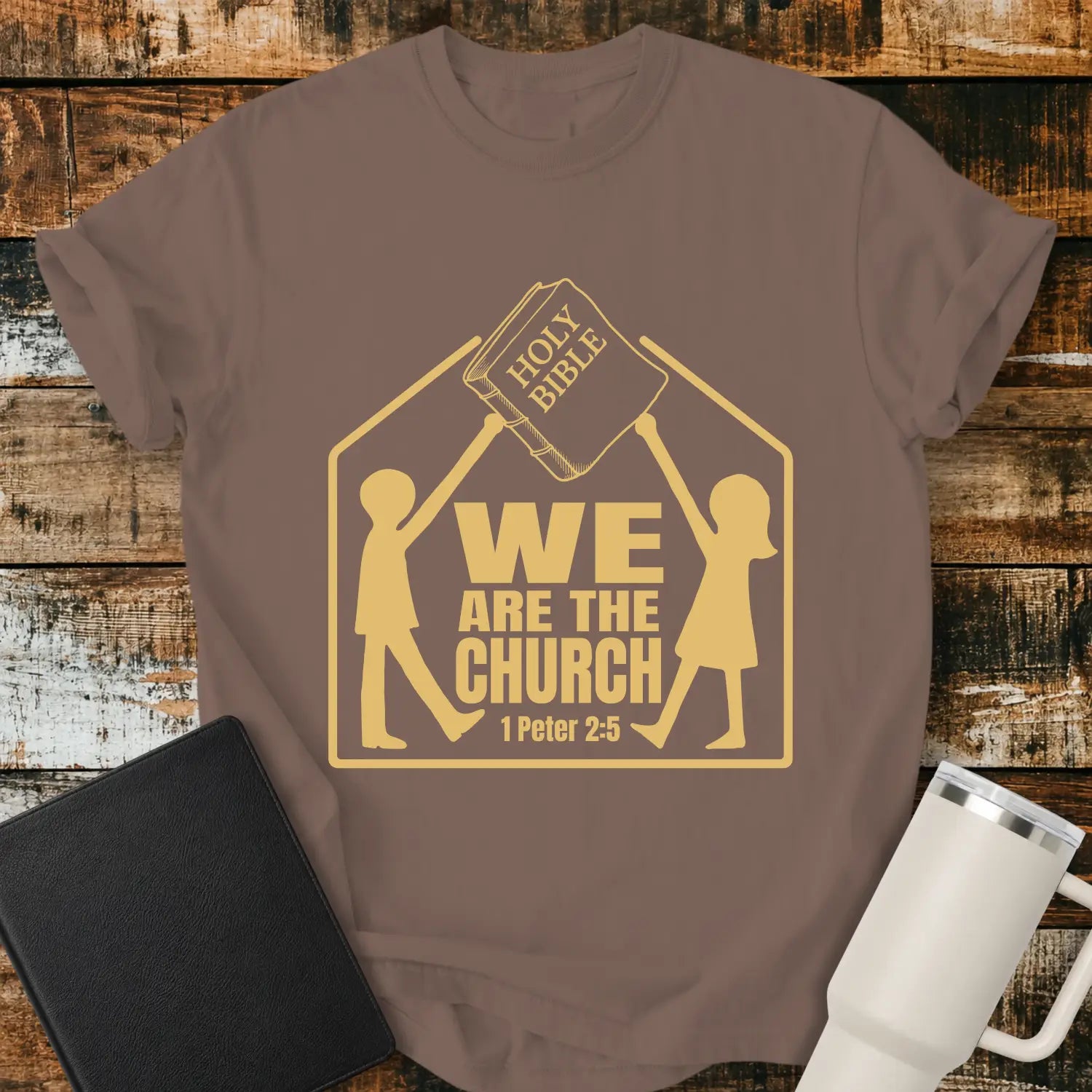 espresso brown graphic T-shirt mockup featuring the phrase “We Are the Church” with a 1 Peter 2:5 reference, showing two figures holding a Holy Bible above a church-shaped outline, styled with a black Bible and white tumbler on a rustic wood background.