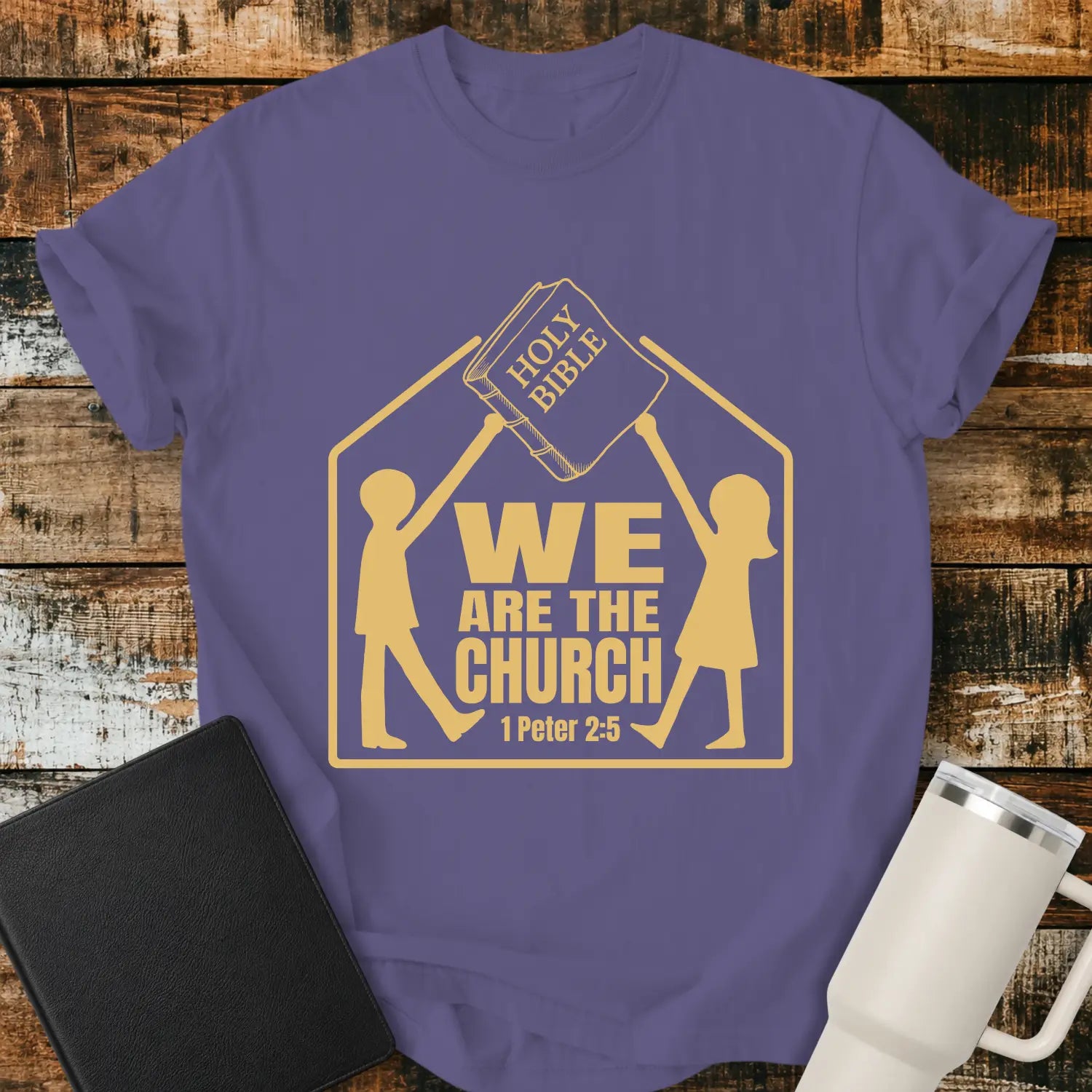 grape graphic T-shirt mockup featuring the phrase “We Are the Church” with a 1 Peter 2:5 reference, showing two figures holding a Holy Bible above a church-shaped outline, styled with a black Bible and white tumbler on a rustic wood background.