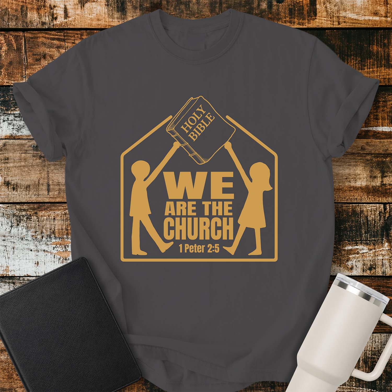 Graphite T-shirt featuring a gold graphic of two children raising a Holy Bible above a house-shaped outline with the message “WE ARE THE CHURCH” and the Bible reference “1 Peter 2:5.” The shirt is displayed on a rustic wood plank background alongside a black Bible and a white insulated tumbler.