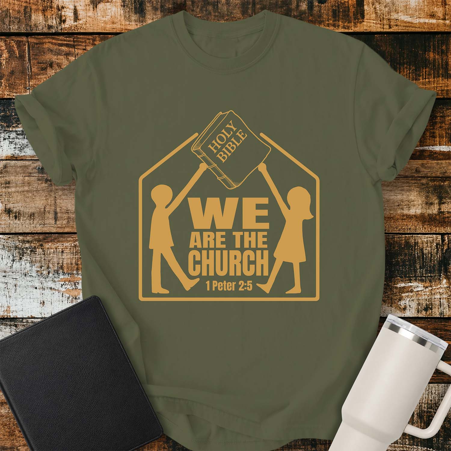 Hemp T-shirt featuring a gold graphic of two children raising a Holy Bible above a house-shaped outline with the message “WE ARE THE CHURCH” and the Bible reference “1 Peter 2:5.” The shirt is displayed on a rustic wood plank background alongside a black Bible and a white insulated tumbler.
