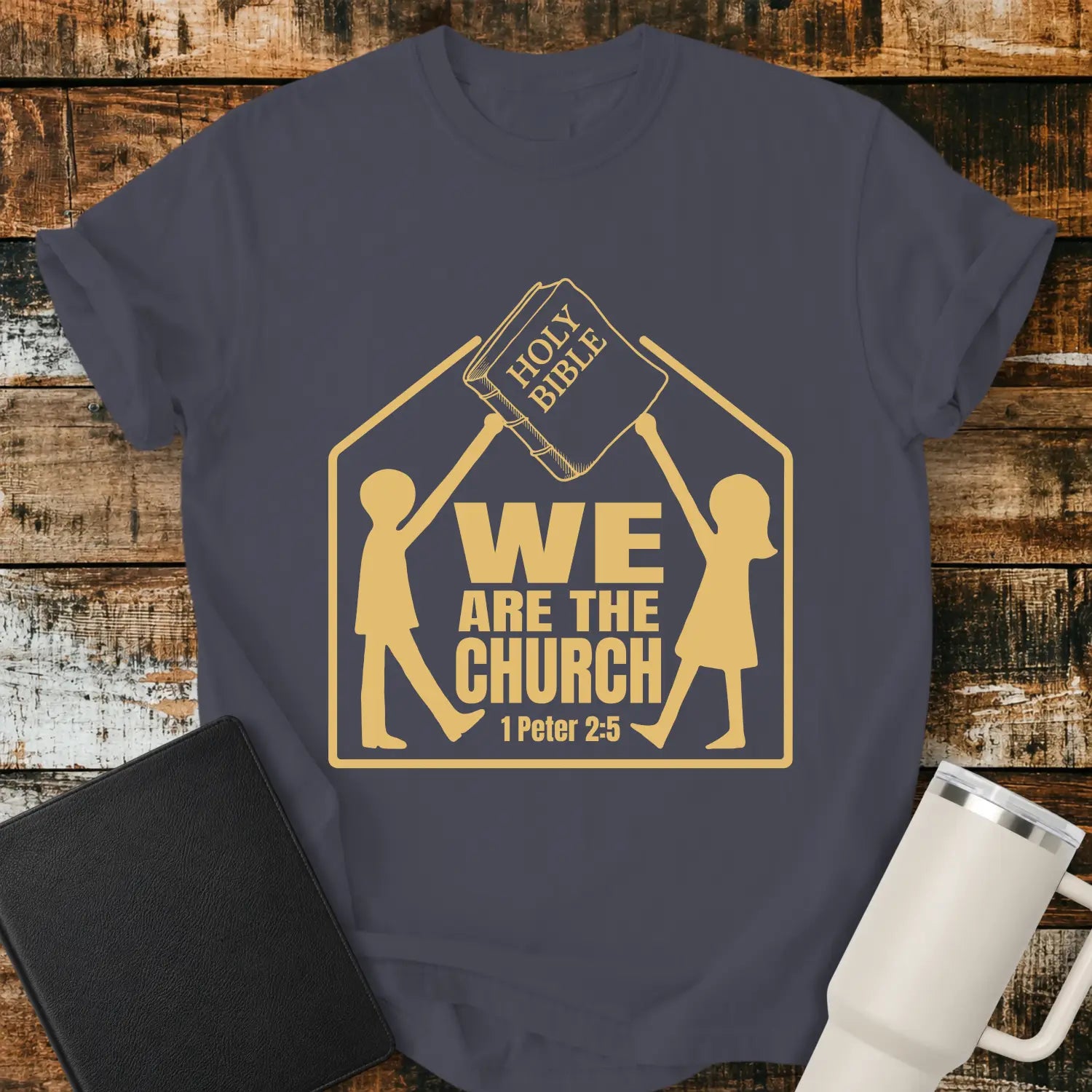 navy blue graphic T-shirt mockup featuring the phrase “We Are the Church” with a 1 Peter 2:5 reference, showing two figures holding a Holy Bible above a church-shaped outline, styled with a black Bible and white tumbler on a rustic wood background.