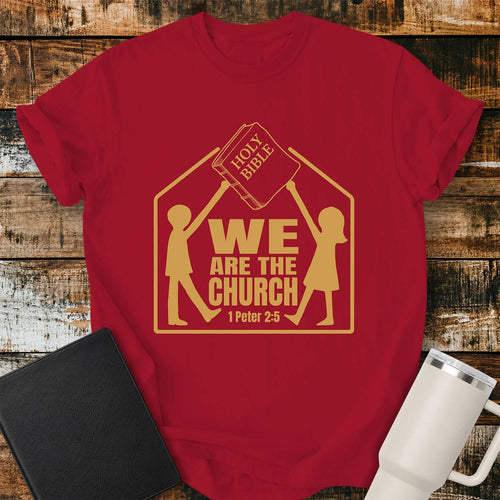 Red T-shirt featuring a gold graphic of two children raising a Holy Bible above a house-shaped outline with the message “WE ARE THE CHURCH” and the Bible reference “1 Peter 2:5.” The shirt is displayed on a rustic wood plank background alongside a black Bible and a white insulated tumbler.