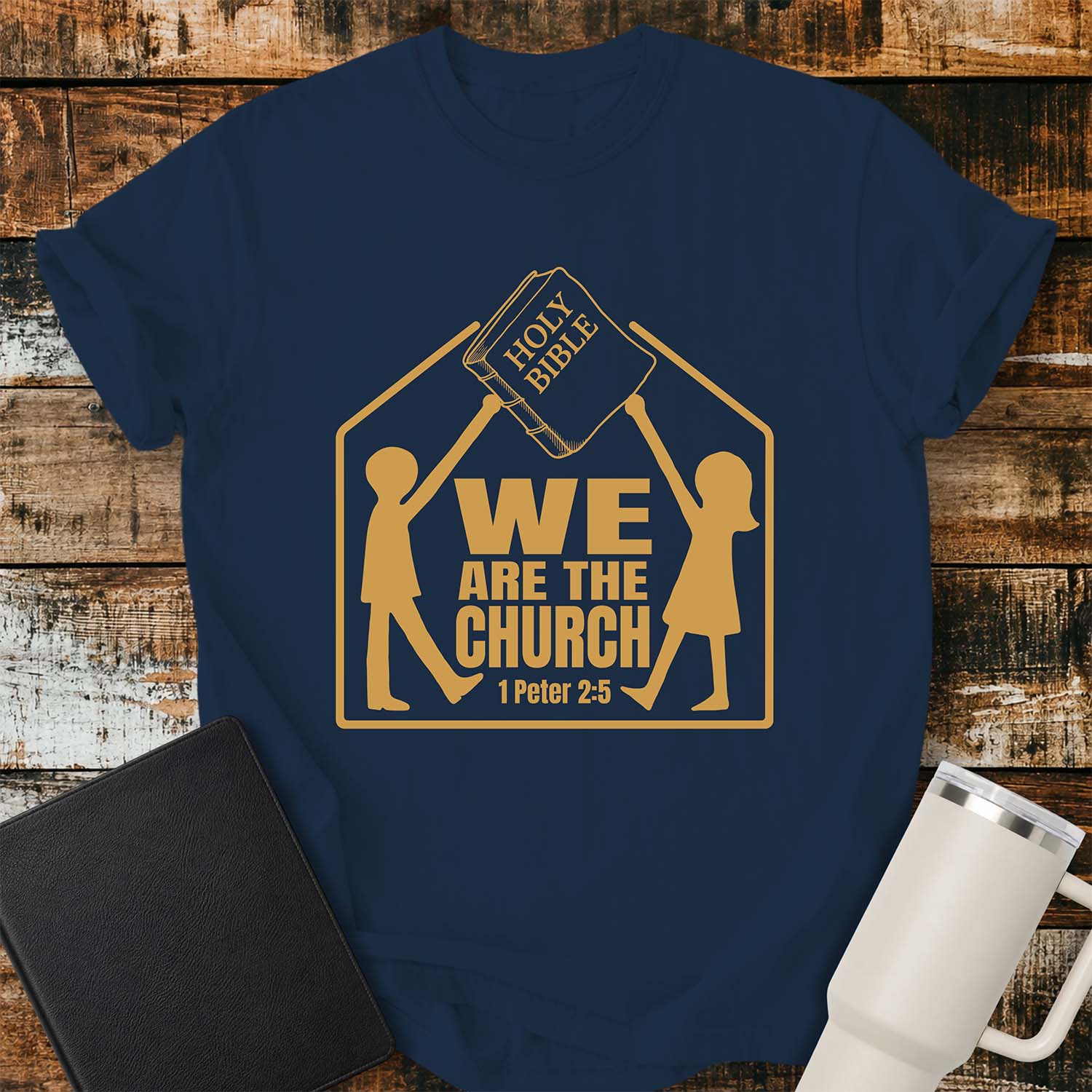 True navy t-shirt featuring a gold graphic of two children raising a Holy Bible above a house-shaped outline with the message “WE ARE THE CHURCH” and the Bible reference “1 Peter 2:5.” The shirt is displayed on a rustic wood plank background alongside a black Bible and a white insulated tumbler.