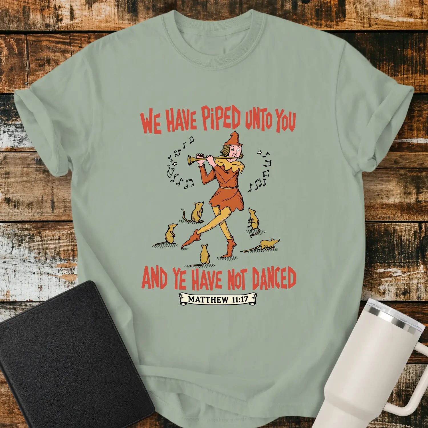 bay green T-shirt mockup featuring a vintage-style illustration of a pipe player surrounded by mice, with the Bible verse phrase “WE HAVE PIPED UNTO YOU AND YE HAVE NOT DANCED” in retro orange lettering and “MATTHEW 11:17” below. The shirt is displayed on a rustic wood background alongside a black Bible and a white insulated tumbler.