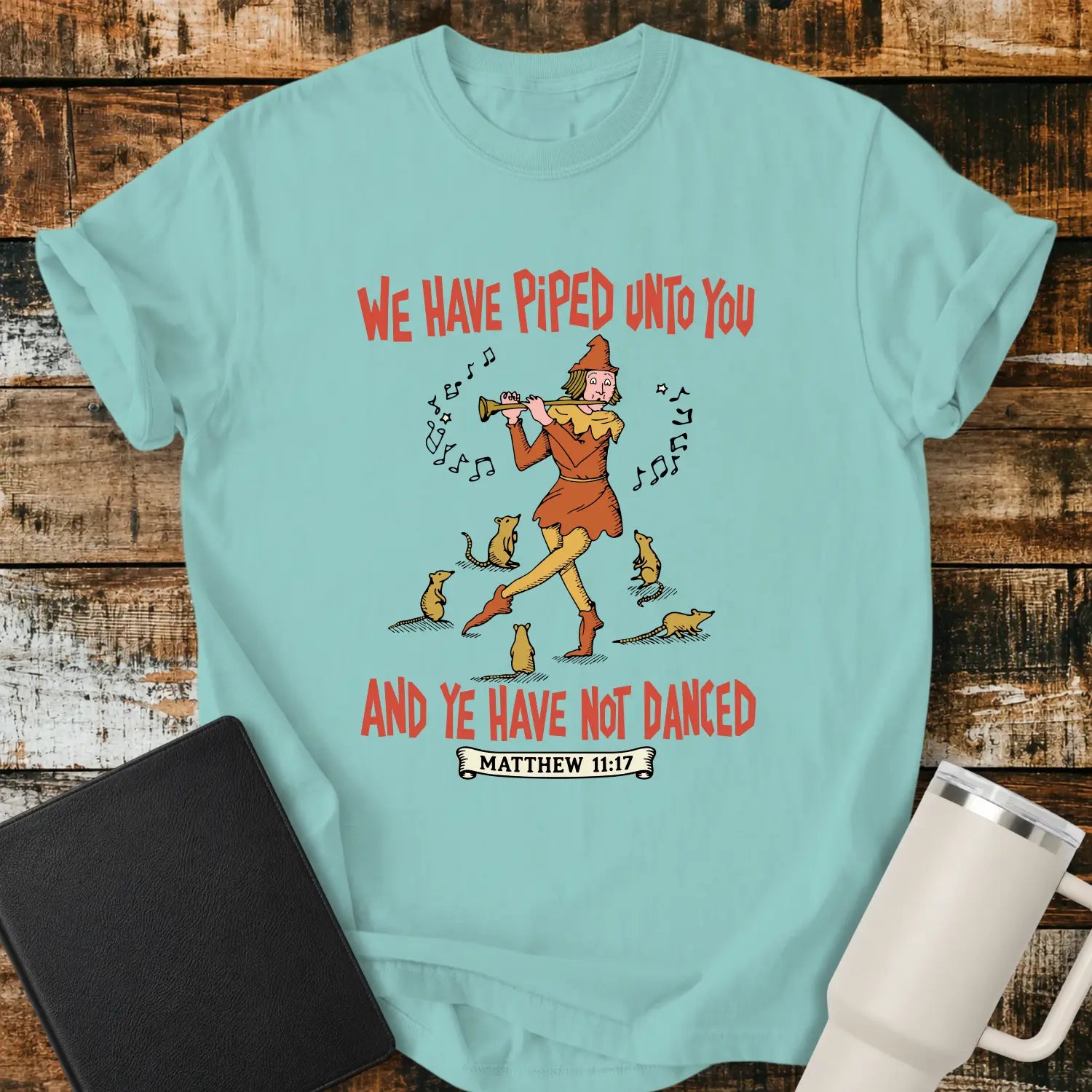 Chalky mint T-shirt mockup featuring a vintage-style illustration of a pipe player surrounded by mice, with the Bible verse phrase “WE HAVE PIPED UNTO YOU AND YE HAVE NOT DANCED” in retro orange lettering and “MATTHEW 11:17” below. The shirt is displayed on a rustic wood background alongside a black Bible and a white insulated tumbler.