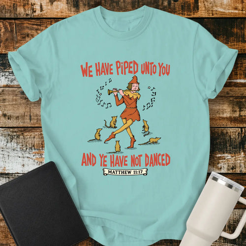 Chalky mint T-shirt mockup featuring a vintage-style illustration of a pipe player surrounded by mice, with the Bible verse phrase “WE HAVE PIPED UNTO YOU AND YE HAVE NOT DANCED” in retro orange lettering and “MATTHEW 11:17” below. The shirt is displayed on a rustic wood background alongside a black Bible and a white insulated tumbler.