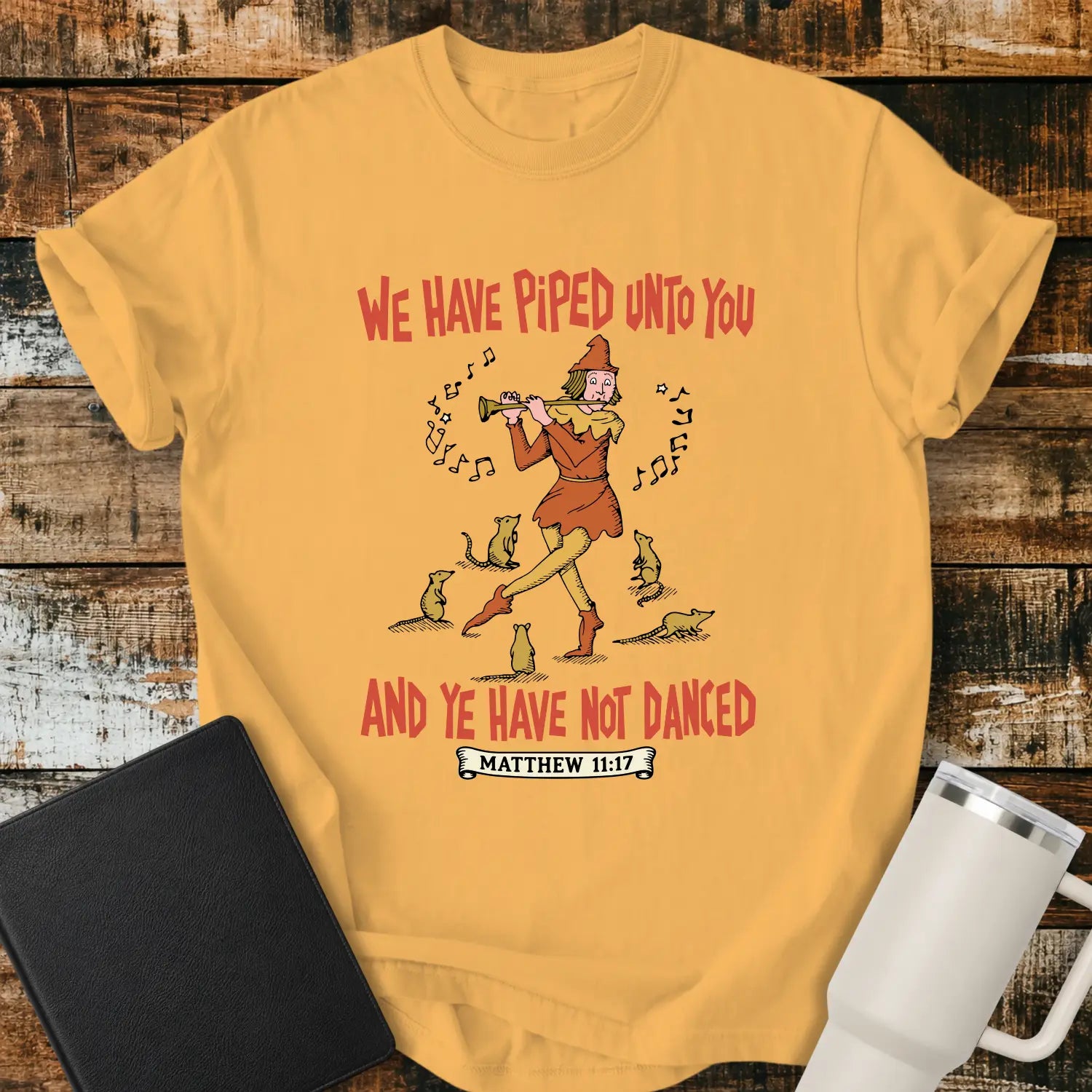 citrus yellow T-shirt mockup featuring a vintage-style illustration of a pipe player surrounded by mice, with the Bible verse phrase “WE HAVE PIPED UNTO YOU AND YE HAVE NOT DANCED” in retro orange lettering and “MATTHEW 11:17” below. The shirt is displayed on a rustic wood background alongside a black Bible and a white insulated tumbler.