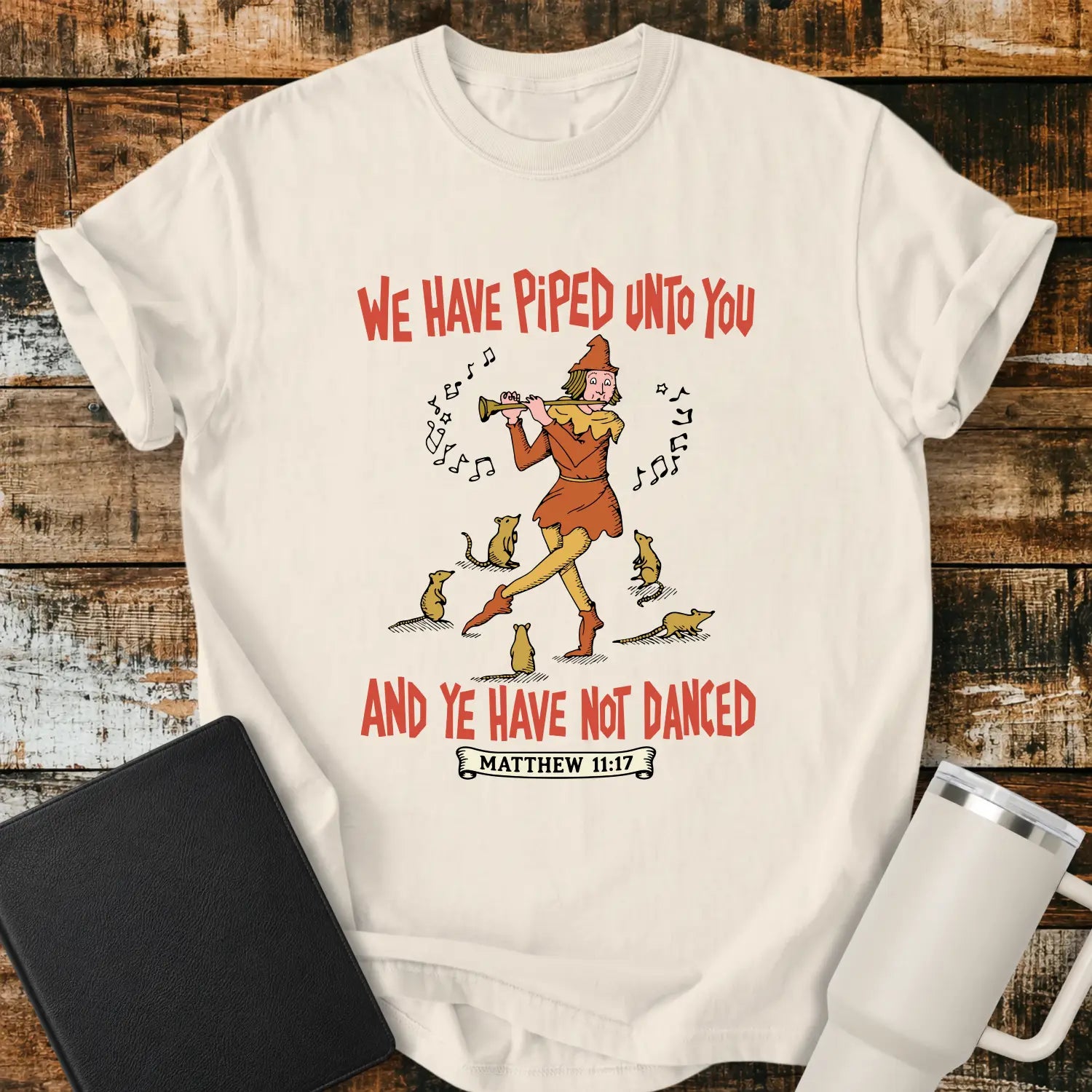 ivory T-shirt mockup featuring a vintage-style illustration of a pipe player surrounded by mice, with the Bible verse phrase “WE HAVE PIPED UNTO YOU AND YE HAVE NOT DANCED” in retro orange lettering and “MATTHEW 11:17” below. The shirt is displayed on a rustic wood background alongside a black Bible and a white insulated tumbler.