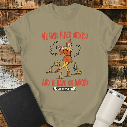 khaki T-shirt mockup featuring a vintage-style illustration of a pipe player surrounded by mice, with the Bible verse phrase “WE HAVE PIPED UNTO YOU AND YE HAVE NOT DANCED” in retro orange lettering and “MATTHEW 11:17” below. The shirt is displayed on a rustic wood background alongside a black Bible and a white insulated tumbler.