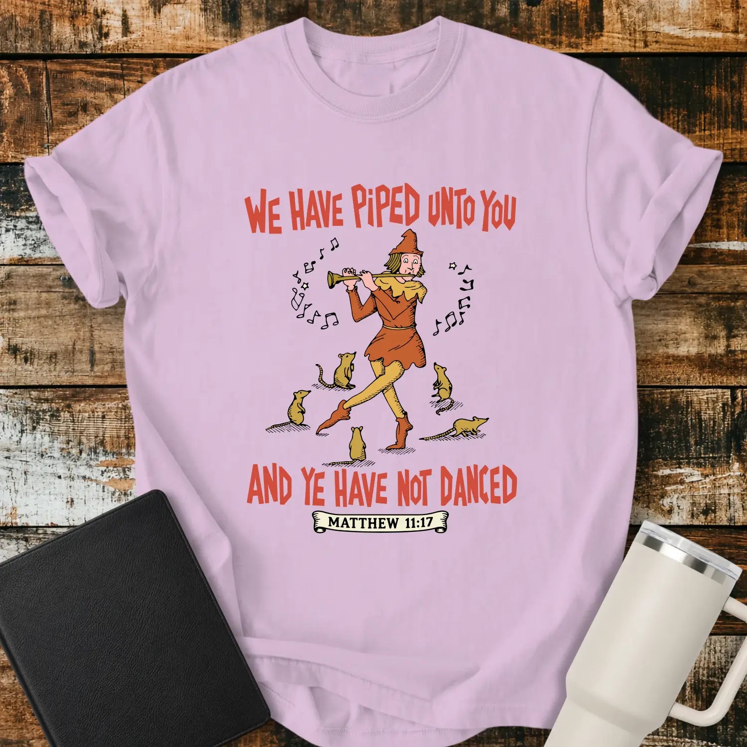 orchid pink T-shirt mockup featuring a vintage-style illustration of a pipe player surrounded by mice, with the Bible verse phrase “WE HAVE PIPED UNTO YOU AND YE HAVE NOT DANCED” in retro orange lettering and “MATTHEW 11:17” below. The shirt is displayed on a rustic wood background alongside a black Bible and a white insulated tumbler.