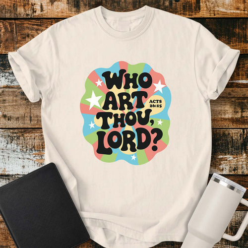 Ivory T-shirt featuring a colorful retro psychedelic graphic with pastel blue, green, pink, and yellow rays, accented by white stars. Bold black bubble lettering reads, “WHO ART THOU, LORD?” with “ACTS 26:15” in smaller text beside the design. The shirt is displayed on a rustic wood plank background with a black Bible on the left and a white insulated tumbler on the right.
