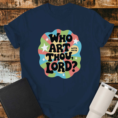 true navy T-shirt featuring a colorful retro psychedelic graphic with pastel blue, green, pink, and yellow rays, accented by white stars. Bold black bubble lettering reads, “WHO ART THOU, LORD?” with “ACTS 26:15” in smaller text beside the design. The shirt is displayed on a rustic wood plank background with a black Bible on the left and a white insulated tumbler on the right.